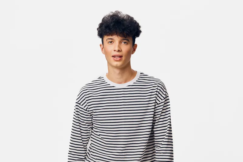 Young man wearing a striped shirt stands against a plain white background, looking directly at the camera with a neutral expression in a studio setting.