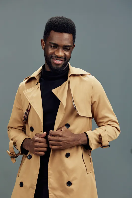 Young African man in a stylish beige trench coat against a muted green background, exuding confidence and a warm smile, perfect for fashion and lifestyle content