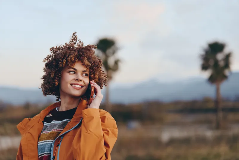 Outdoor woman lifestyle portrait, smiling young woman with curly hair in an orange jacket, gently touching hair outdoors in a rural field under warm evening light, softly blurred trees