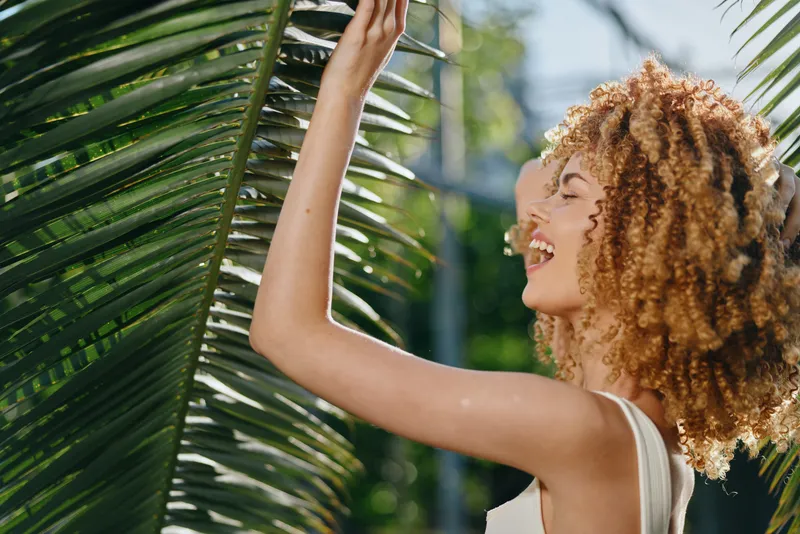 Smiling woman with curly hair enjoys nature under sunlight near tropical palm leaves in warm outdoor setting with green blurred background and natural light.
