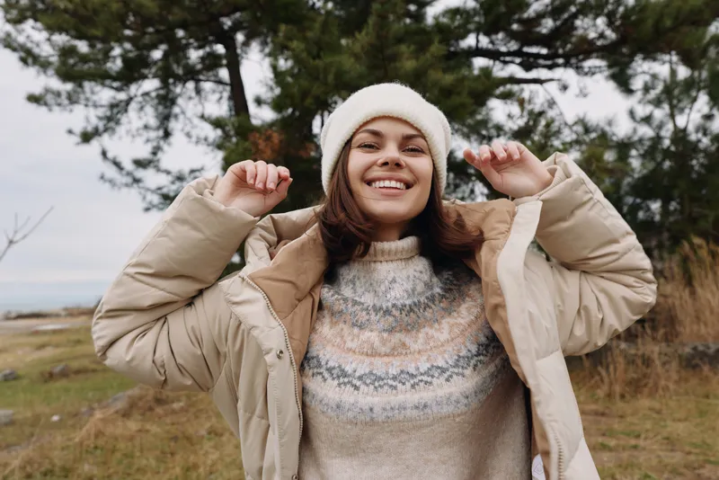 Woman outdoors in a cozy sweater and puffy jacket, smiling in a natural landscape with trees and an open field, conveying warmth, comfort, and casual winter fashion in a candid lifestyle shot.