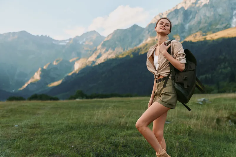 Young woman enjoys outdoor hiking in a mountain meadow, wearing a shirt and shorts with a backpack, embracing nature and adventure in bright daylight