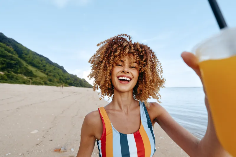 A joyful young woman with curly hair wears a colorful striped swimsuit and raises a beverage on a sunny beach, conveying a carefree vacation moment.