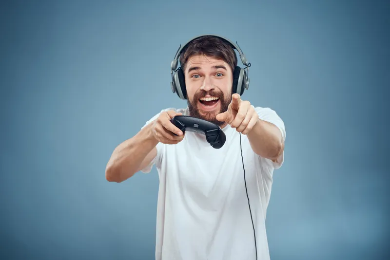 Мужчина в наушниках с контроллером в руках играет на игровой консоли, образ жизни, эмоции, синий фон, белая футболка