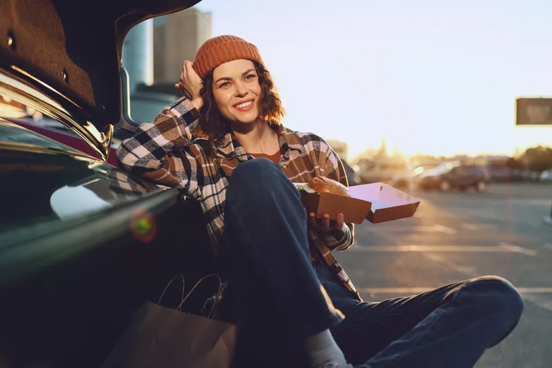 Woman by car in parking lot smiles with takeout box, beanie and casual outfit in goldenhour glow; authenticity and candid lifestyle moment conveying mindful living and emotional storytelling.