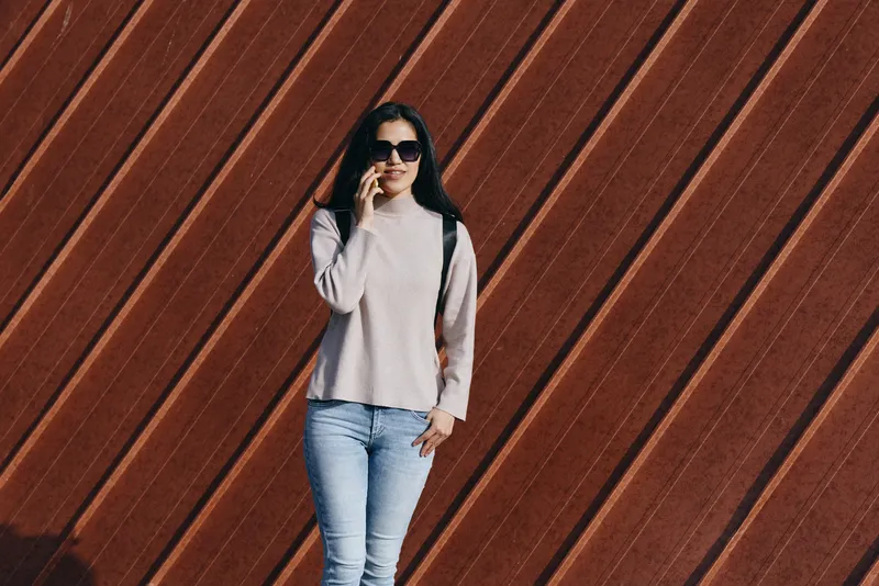 Woman in casual fashion poses against a diagonal wooden wall, wearing sunglasses, a light sweater and jeans with a backpack, showcasing an urban street style moment.