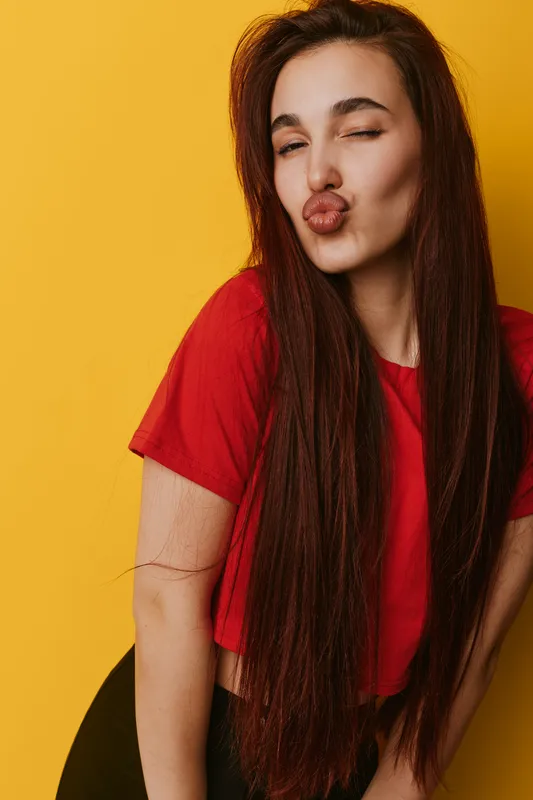 Young woman with long hair wearing red shirt posing with kiss face expression against yellow background. Beauty, fashion and lifestyle portrait with casual style and playful mood.