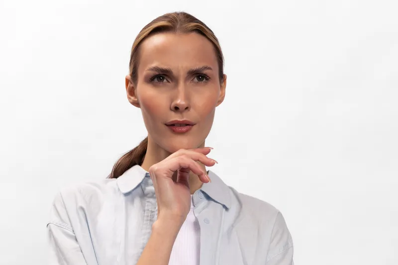 Confused young woman with light skin and tied back hair looks puzzled, holding her chin. Isolated on white background. Concept for doubt, thinking, and uncertainty.
