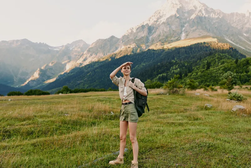 Young woman hiking in a bright meadow beside majestic mountains, carrying a backpack, casual outdoor attire, capturing a spontaneous travel moment in nature