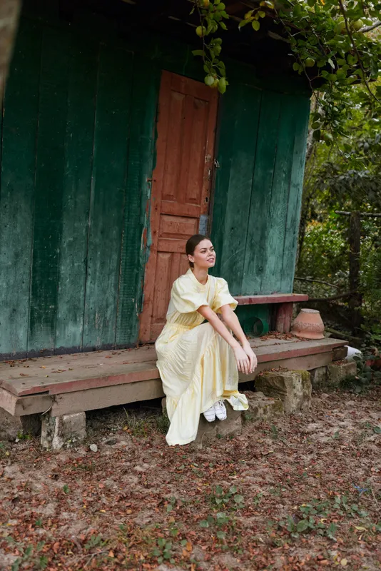A woman in a yellow dress sits on a wooden bench outside a green door, surrounded by trees. A calm, reflective moment in a rustic outdoor setting with soft natural light.
