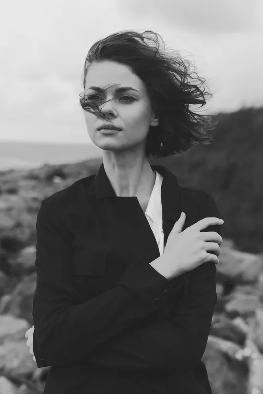 Woman standing on a windy coastline in a dark jacket, wind tousling hair, a contemplative mood in a monochrome scene for fashion and nature