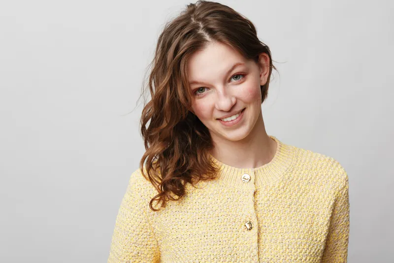 Smiling young woman with curly brown hair wearing yellow knitted sweater looks at camera on grey background. Concept of happiness, casual style, and natural beauty portrait.
