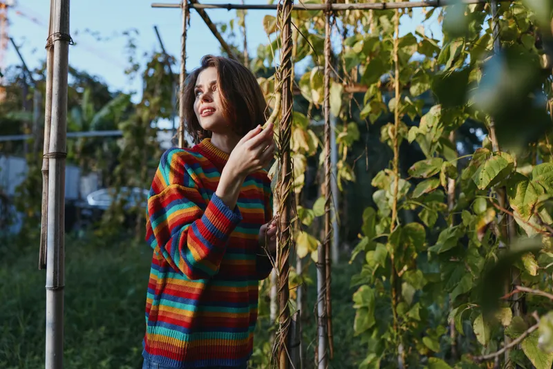 Woman in colorful striped sweater in garden among climbing plants on trellis and vines, outdoor portrait with gentle smile, sunlight and lush green foliage background.