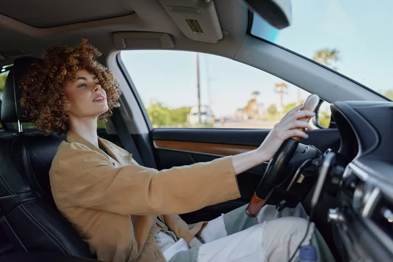 A woman driver sits behind the wheel in a tan jacket, focused on the road. Bright daylight filters through the car window, highlighting a stylish, casual lifestyle moment.