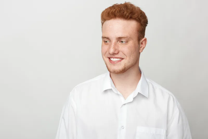 Smiling young man with red hair wearing white shirt looks sideways on light background. Casual portrait of happy male model with natural expression and clean studio lighting.