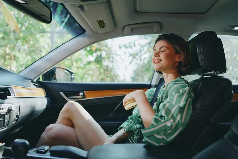 A woman sits in a car enjoying a sandwich, smiling contentedly as daylight fills the cabin.