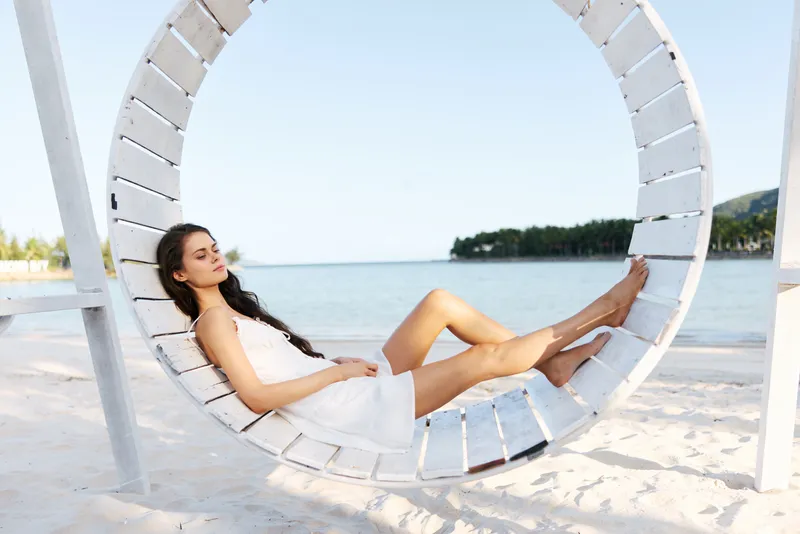 Relaxed woman in white dress sitting on circular swing by the beach under clear sky, embodying calmness and summer vibes with natural scenery.
