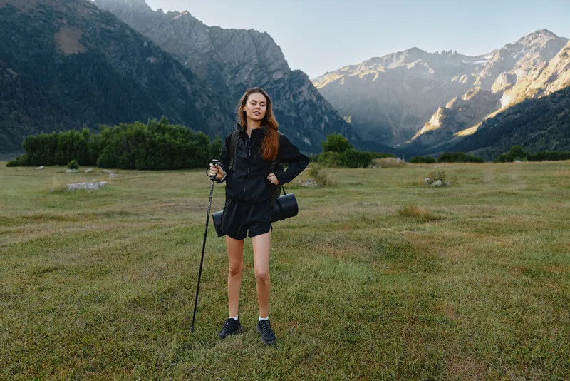 A confident young woman stands in a wide grassy valley with towering rugged mountains behind, gripping a trekking pole and wearing outdoor gear. She smiles, ready for adventure.