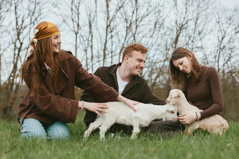 A joyful trio outdoors with a playful goat kid, as friends gently pat the animals. Casual lifestyle scene highlighting bonding, care, and rural simplicity.
