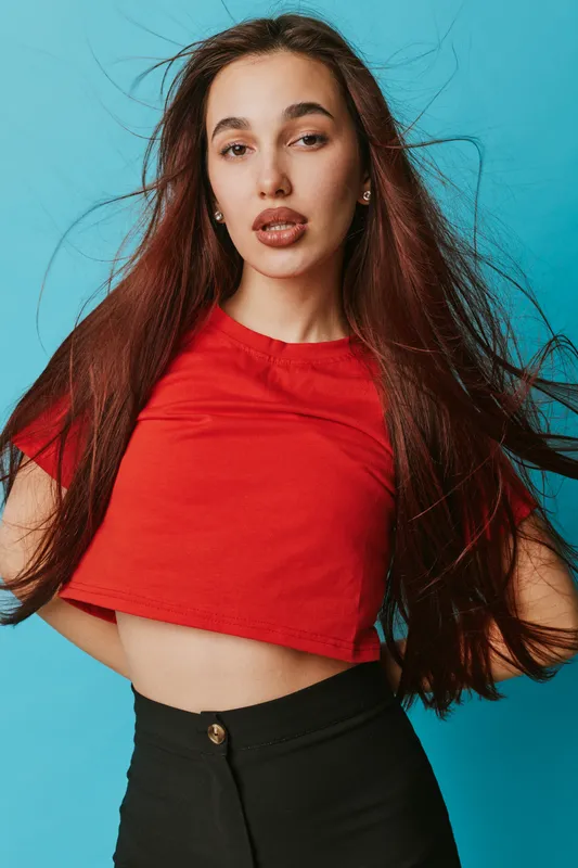 Woman with long hair wearing red crop top and black pants posing against blue background. Fashion portrait with natural makeup and confident expression for style and beauty concept.