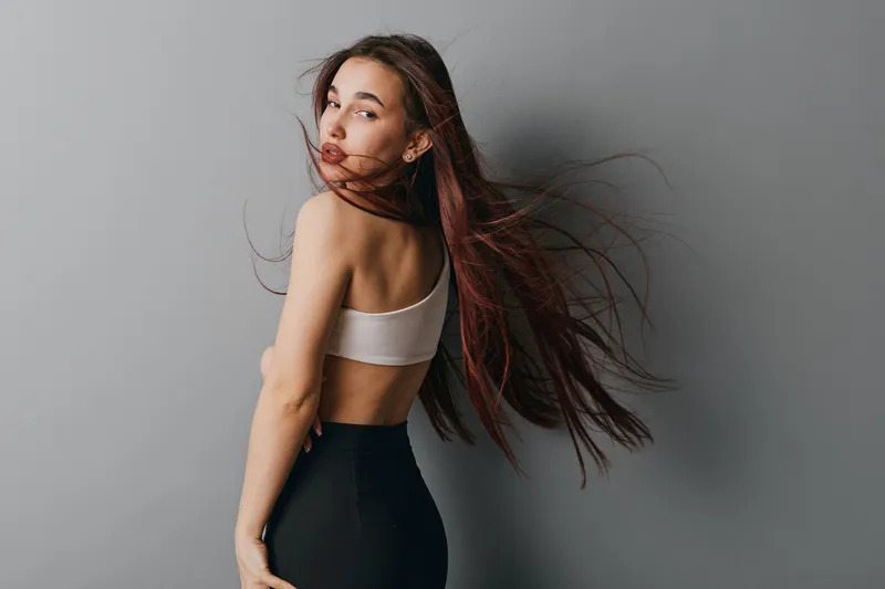 Young woman with long hair wearing white top and black pants posing against gray background. Female model with flowing hair and casual outfit looking over shoulder with confident expression.