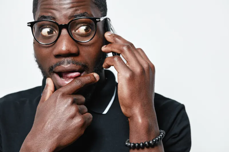 African American man with dark skin and glasses talking on smartphone with surprised expression wearing black polo shirt and bracelet on white background.