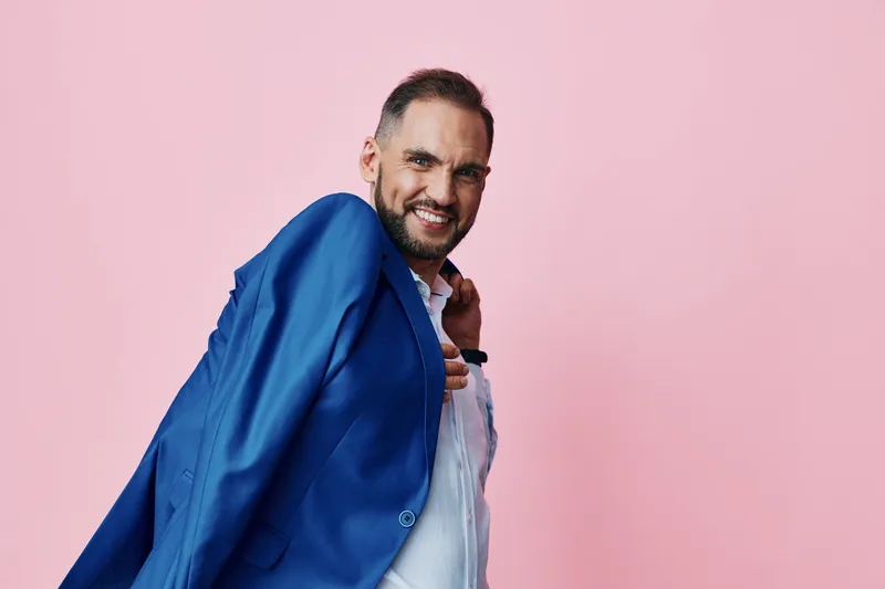 A professional man on a solid pink background smiles confidently, draping a blue suit over his shoulder. The isolated color setting conveys ambition and readiness for business.