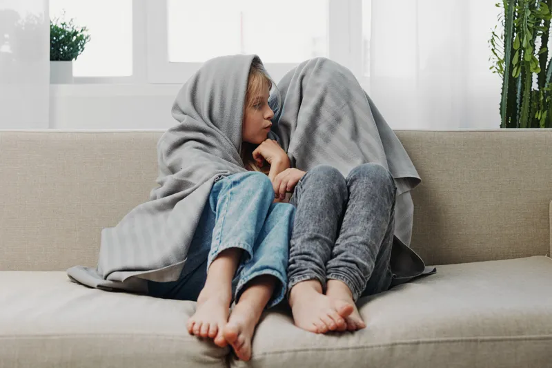 Two children sitting closely on a beige sofa wrapped in a grey blanket, barefoot and thoughtful in a bright living room with plants and natural light. Concept for comfort and childhood.