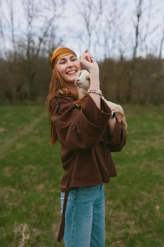 Smiling woman with a goat kid outdoors, wearing a brown jacket and yellow headband, enjoying a playful moment in nature. Lifestyle scene highlighting bonding, wellness, and casual fashion in soft
