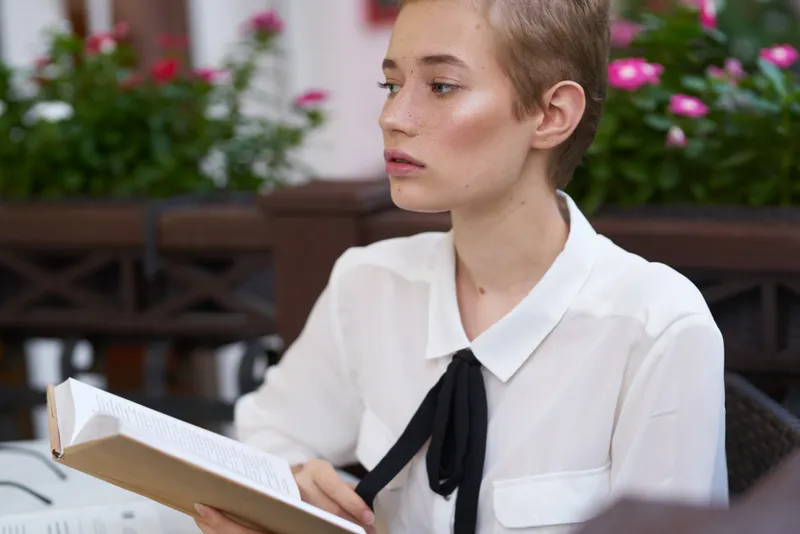 Элегантная женщина в рубашке с галстуком держит книгу в руке, на фоне кафе, улица, деревья, цветы