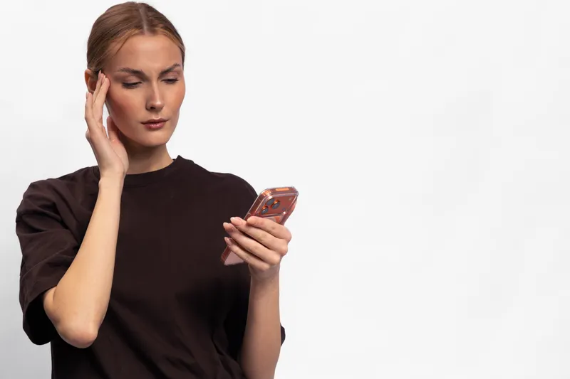 Young woman in dark shirt looks confused and worried while checking smartphone, isolated on white background. Concept for stress, communication problems, and digital frustration.