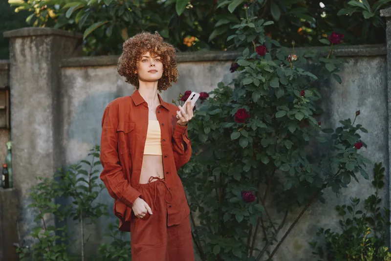 Woman outdoor in rust red outfit stands beside a garden wall, looking at her phone with a relaxed smile, warm light, gentle breeze, lush greenery and blossoms in natural colors