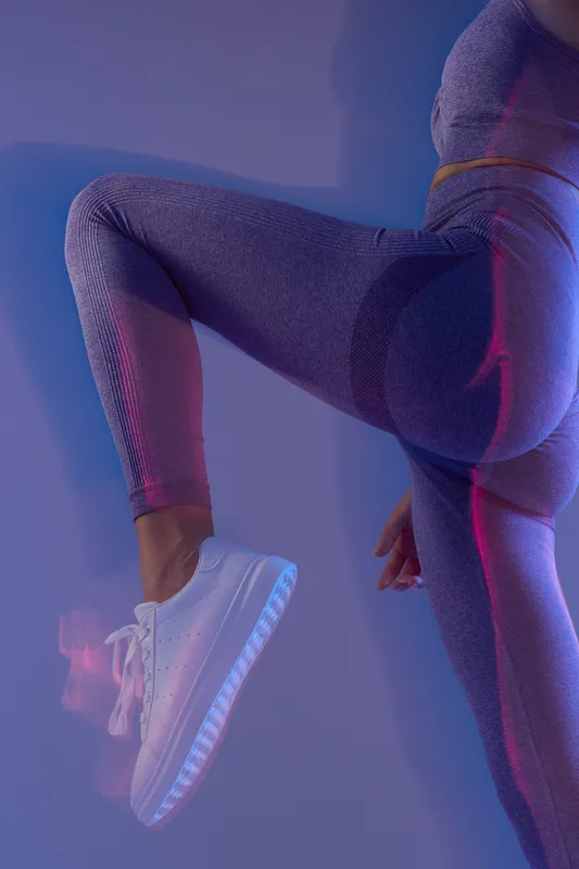 Active woman sport fitness in textured purple leggings and white sneakers, performing a leg raise exercise under colorful lighting, showcasing strength and flexibility.