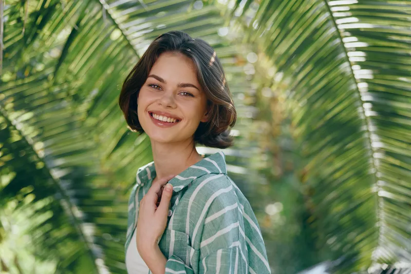 Smiling young woman stands among tropical palm leaves, wearing a striped green shirt, bathed in bright outdoor light.