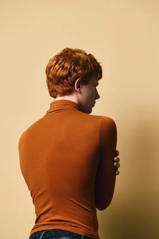 Young man with red hair wearing orange long sleeve sweater standing back to camera posing against light beige background. Studio portrait of person isolated.