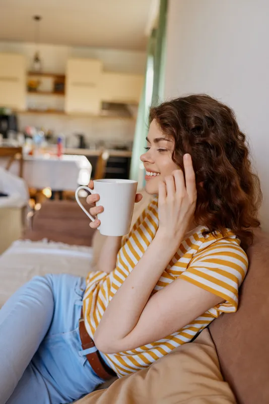 A woman in a striped tshirt sits on a sofa at home, smiling while holding a mug and talking on the phone. A cozy, casual lifestyle concept focused on warmth and everyday comfort.