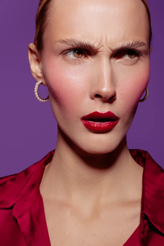 Close-up of a serious woman with red lipstick and hoop earrings wearing a red blouse against a purple background. Concept of fashion, beauty, and intense emotion.