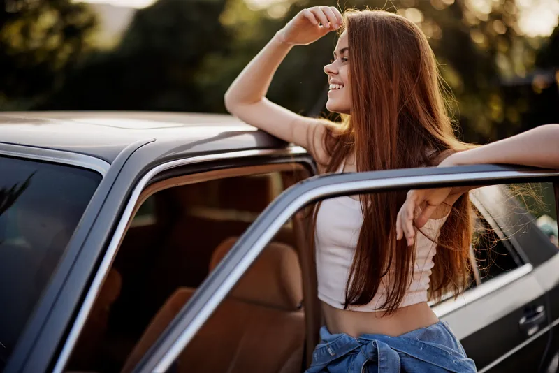 Young woman enjoying a sunny day, leaning on a vintage car with a joyful expression, highlighting carefree summer vibes and fashion style.