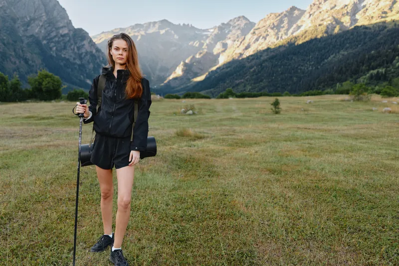 A young woman stands in a wide mountain meadow, outfitted in hiking gear with a trekking pole and camera bag, preparing for an alpine trek in pristine nature.