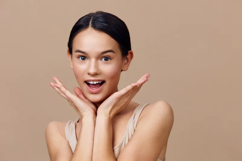 Cheerful young woman expressing surprise with hands on cheeks, wearing a neutral top against a soft beige background, ideal for beauty and emotion focused projects