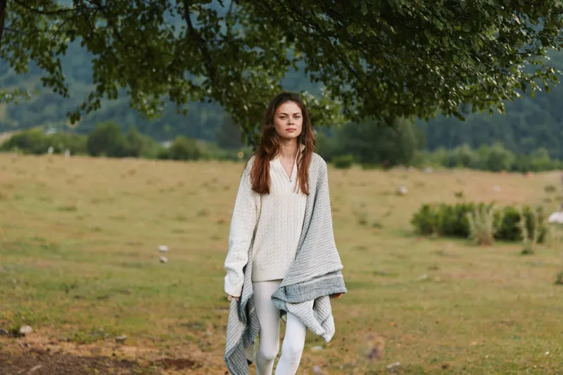 Young woman with long brown hair walks in an open field under a shade tree, wearing a light knit sweater and a soft blanket draped over her shoulders.