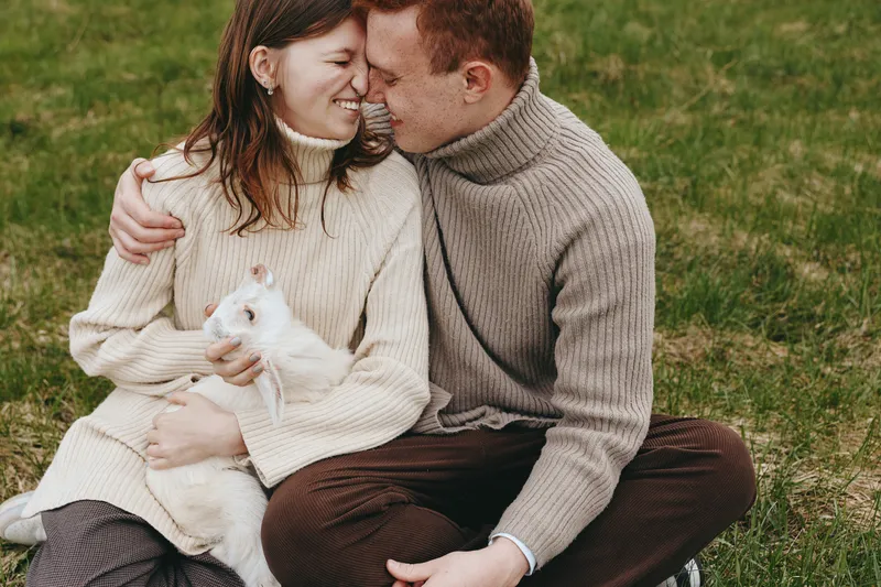 A cozy couple sits on grass, cuddling a small goat kid in warm sweaters, capturing a heartfelt lifestyle moment of affection and companionship.