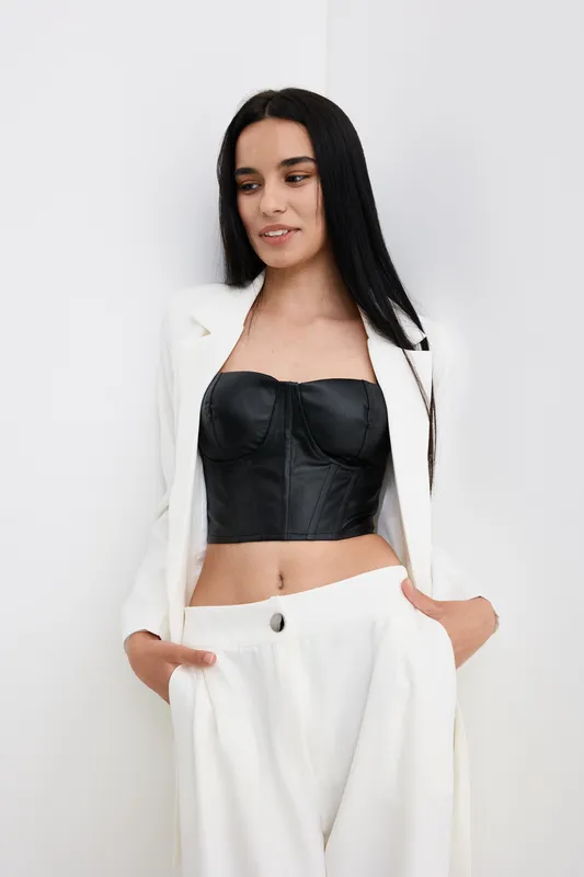 Young woman with long black hair wearing white suit and black leather corset top posing with hands in pockets against plain white background.
