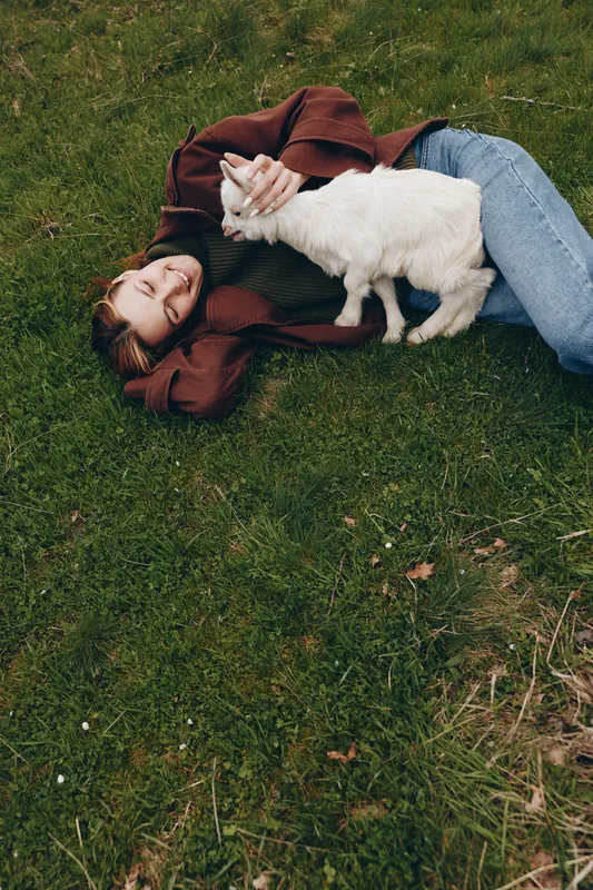 Cheerful woman outdoors, lying on grass, goat kid nuzzles her hand, relaxed pose, sunlit scene, natural colors, gentle breeze, countryside mood, authentic lifestyle moment, joyful connection