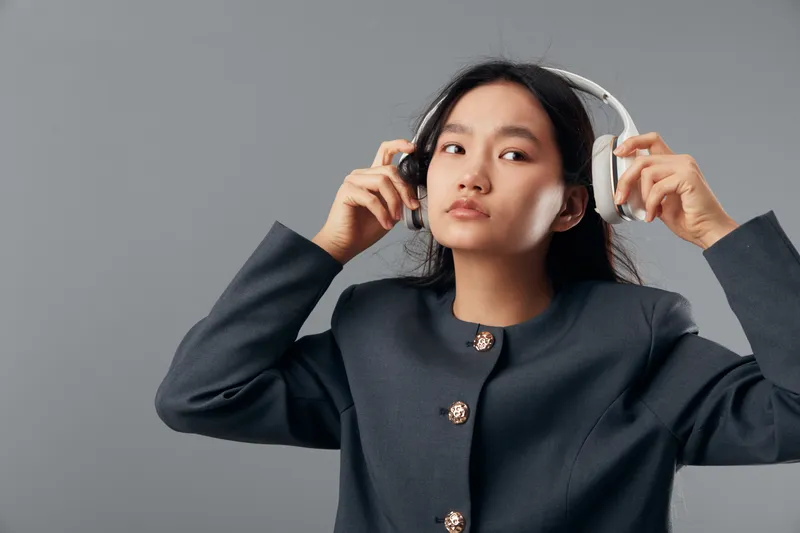 Young woman with long black hair wearing dark jacket adjusts white headphones against grey background for music listening and audio enjoyment in studio or indoor space.