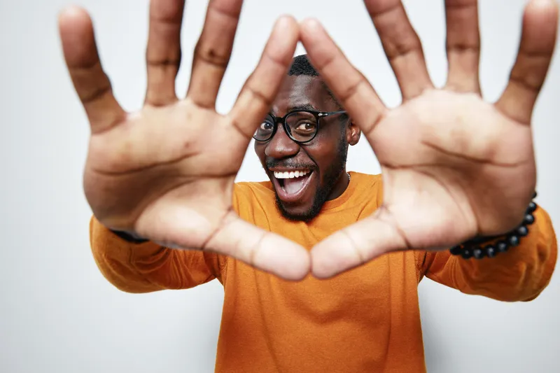 African American man with dark skin wearing glasses and orange sweater smiling and showing hands close to camera in studio. Happy male portrait with joyful expression and casual style.
