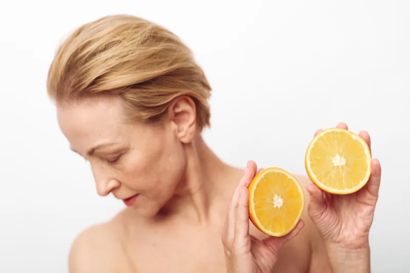 Blonde woman with bare shoulders holds two halves of an orange close to her face, looking down thoughtfully. Bright white background highlights fresh citrus and soft skin texture.
