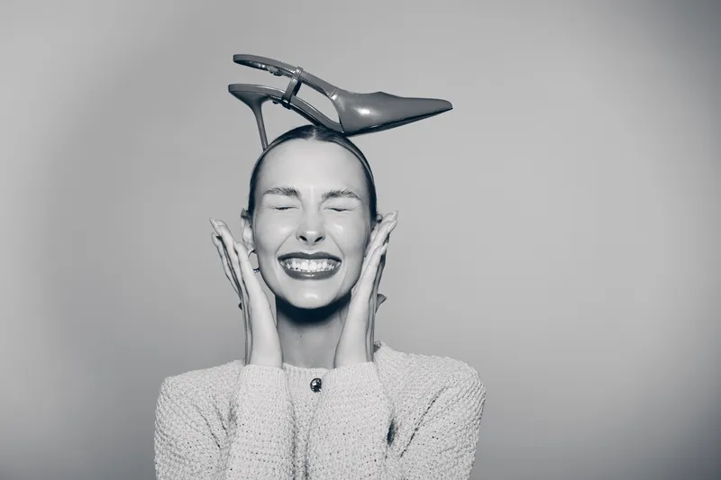 Smiling Caucasian woman with light skin holding high heel shoe on head with closed eyes and joyful expression in studio.