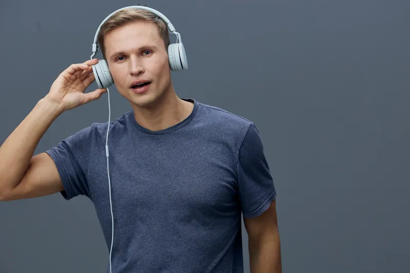 Young man wearing blue t-shirt and headphones listening to music, looking surprised or confused, standing against plain grey background, studio portrait, people lifestyle concept