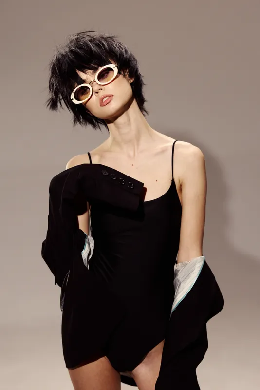 Stylish woman in black outfit wearing sunglasses with short messy hair and a neutral gray background